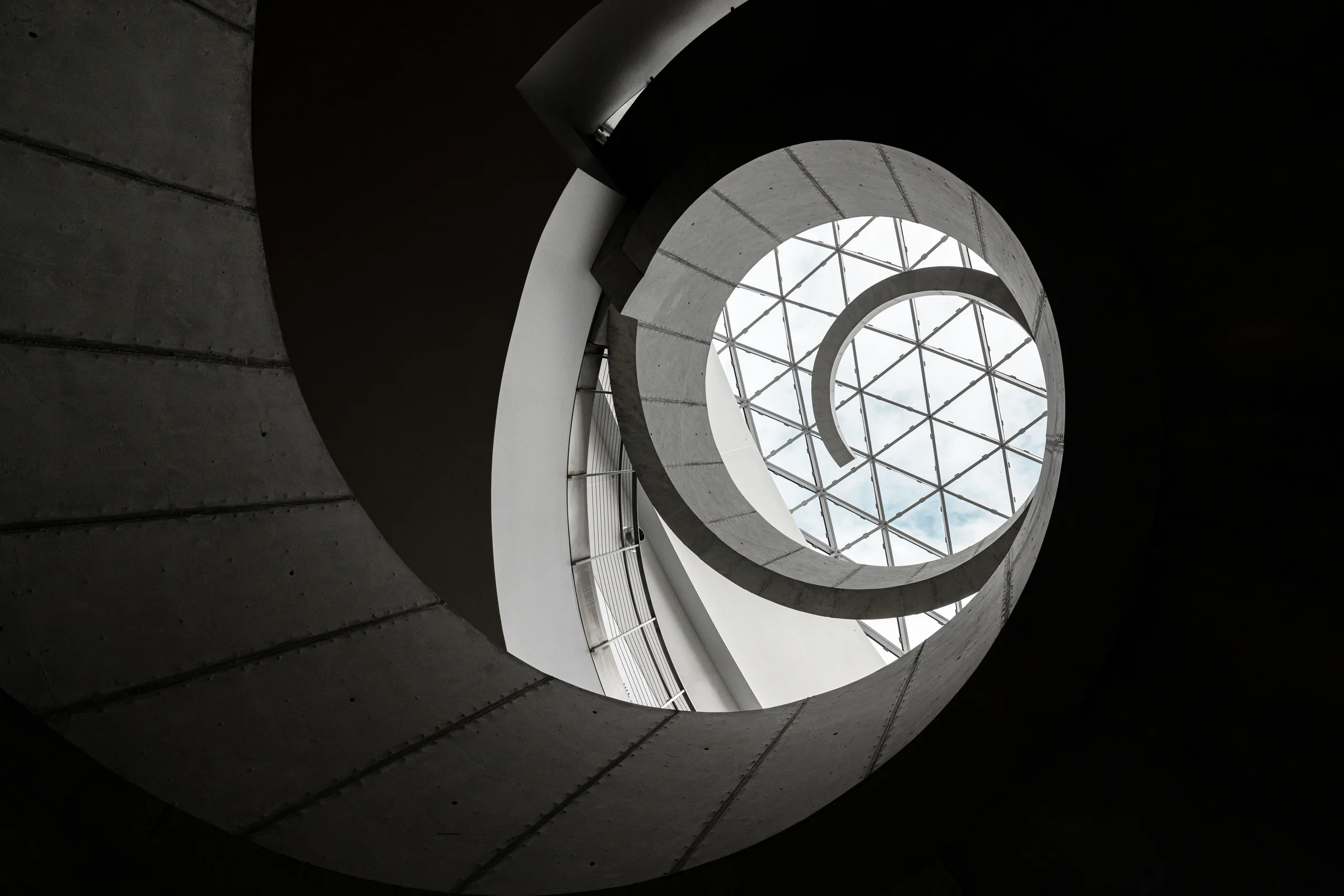 Black-and-white photograph looking up a spiral staircase toward a triangular glass skylight, by ARCHTRAITS